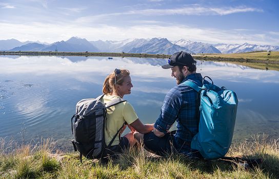 Zwei Menschen sitzen mit Rucksäcken an einem See und genießen die Aussicht auf schneebedeckte Berge und klaren blauen Himmel. Sie lachen und entspannen sich.