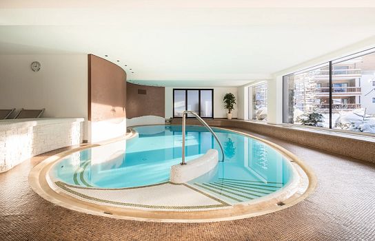 Ein eleganter, ovaler Innenpool mit Stufen, hell erleuchtet durch große Fenster. Die stilvolle Einrichtung umfasst Holz- und Fliesenakzente sowie schlichte Pflanzen.