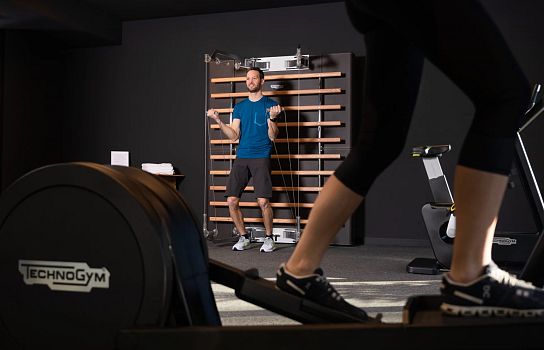 Eine Person in Sportkleidung steht vor einer Kletterwand in einem Fitnessraum. Im Vordergrund läuft jemand auf einem Crosstrainer.