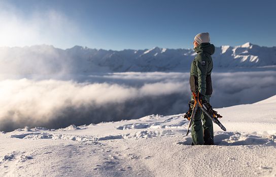 Person in Winterkleidung steht auf verschneitem Berg und blickt in die Ferne. Die Sonne scheint über einem Meer aus Wolken und schneebedeckten Gipfeln.