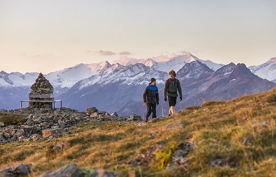 Zwei Wanderer gehen auf einem Bergpfad in den Alpen, umgeben von schneebedeckten Gipfeln. Ein Steinhaufen markiert den Weg, der Himmel ist rosa gefärbt.