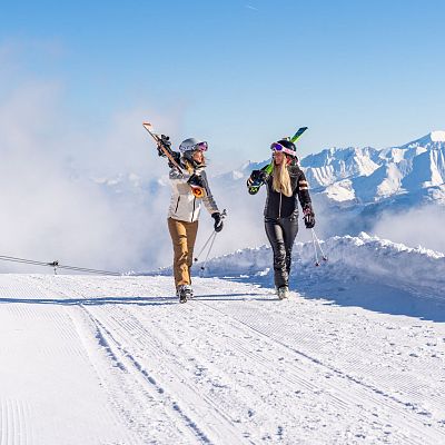 Zwei Menschen gehen auf einer schneebedeckten Piste mit Skiern in den Händen. Im Hintergrund sind verschneite Berge und ein blauer Himmel zu sehen.