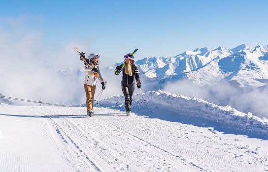 Zwei Menschen gehen auf einer schneebedeckten Piste mit Skiern in den Händen. Im Hintergrund sind verschneite Berge und ein blauer Himmel zu sehen.
