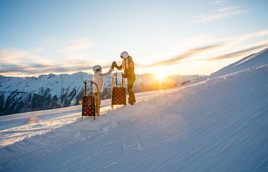 Zwei Personen in Winterkleidung stehen auf einem verschneiten Berg mit Schlitten. Im Hintergrund sind schneebedeckte Berge und der Sonnenuntergang zu sehen.