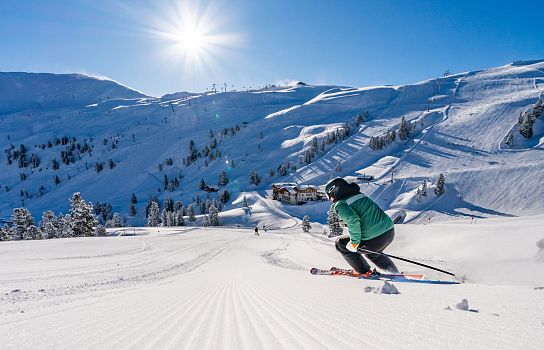 Ein Skifahrer in grüner Jacke fährt eine frisch präparierte Piste hinunter, umgeben von schneebedeckten Bergen und strahlendem Sonnenschein.