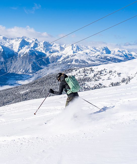 Ein Skifahrer fährt einen schneebedeckten Hang hinab, umgeben von majestätischen, schneebedeckten Bergen. Im Hintergrund sind Skilifte und klare blaue Himmel zu sehen.